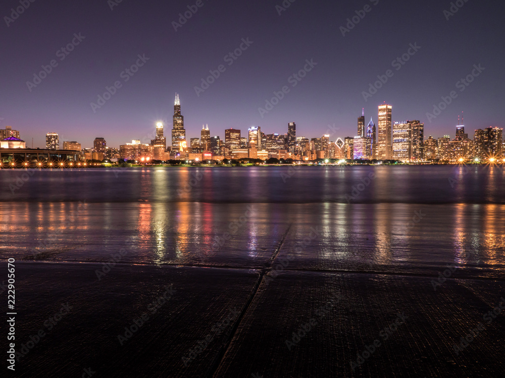 Obraz premium Beautiful long exposure Chicago night skyline photo with colorful red, green, purple, blue, pink, orange, and yellow building lights and water reflections on Lake Michigan and shoreline in foreground