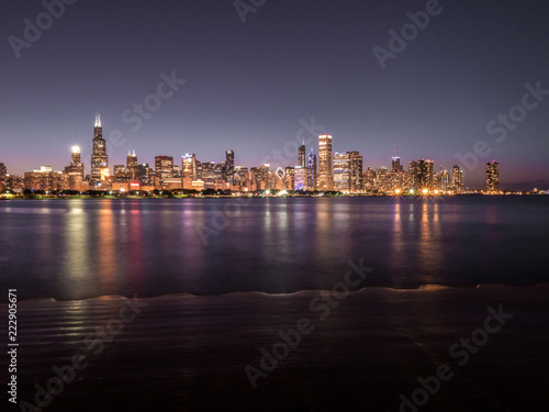 Beautiful long exposure Chicago night skyline photo with colorful red, green, purple, blue, pink, orange, and yellow building lights and water reflections on Lake Michigan and shoreline in foreground