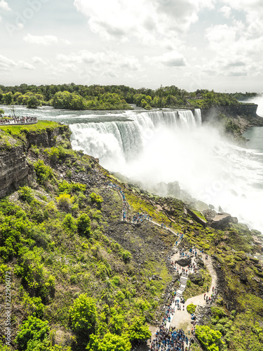 View of the powerful and dramatic American Falls waterfall in Niagara New York with white fluffy clouds above and bright green vegetation and trees growing along the river banks
