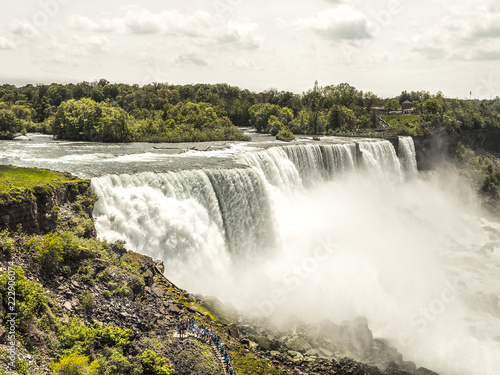 View of the powerful and dramatic American Falls waterfall in Niagara New York with white fluffy clouds above and bright green vegetation and trees growing along the river banks