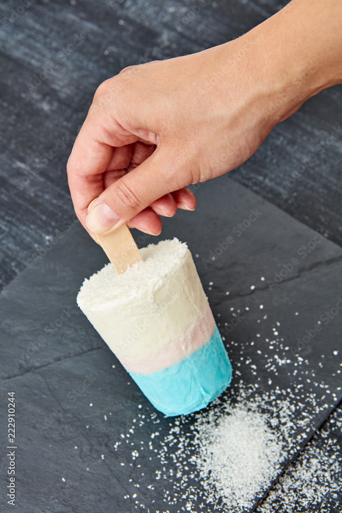 Φωτογραφία A woman's hand holds a coconut ice cream on a stick on a black slate board, with a coconut crumb on a dark concrete background with a copy of the space
