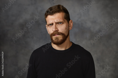 Studio shot of emotional European young male with fuzzy beard frowning, having displeased facial expression, being mad with his children for bad behavior, demonstrating annoyance and irritation