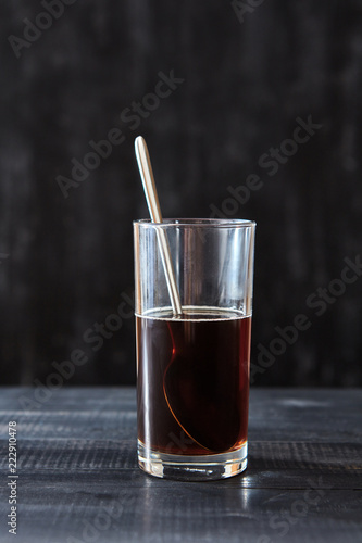 A transparent glass with coffee and a spoon on a black wooden background with copy space.