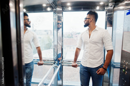 Stylish tall arabian man model in white shirt, jeans and sunglasses posed at elevator inside.