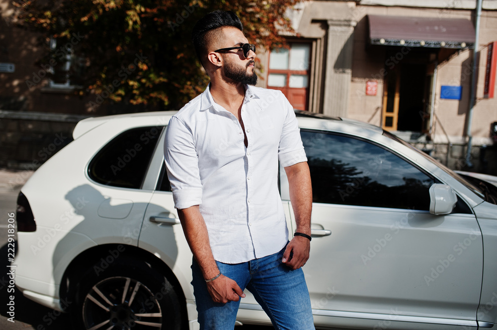 Stylish tall arabian man model in white shirt, jeans and sunglasses posed at street of city. Beard rich attractive arab guy against white suv car. Stylish tall arabian man model in white shirt, jeans and sunglasses posed at street of city. Beard rich attractive arab guy against white suv car.