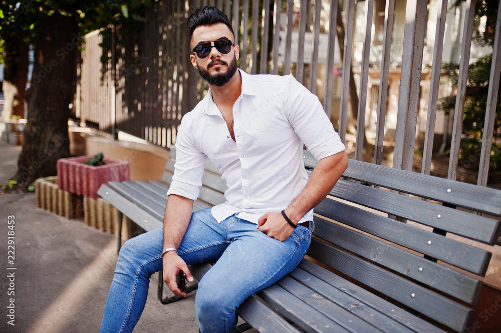 Fototapeta premium Stylish tall arabian man model in white shirt, jeans and sunglasses posed at street of city. Beard attractive arab guy sitting on bench.