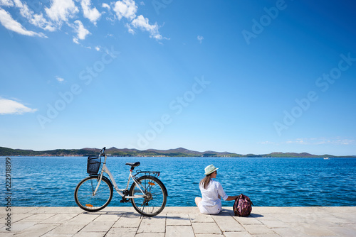 Back view of young woman in white closing, sunhat and sunglasses sitting at bicycle on stony sidewalk under clear blue sky on sparkling clear water background. Tourism and vacations concept.
