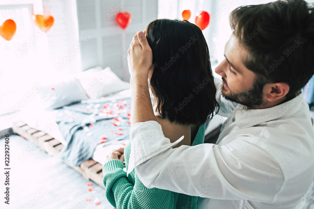 Romantic surprise for your beloved. A handsome man hugs and closes eyes to his girlfriend or his wife making her a romantic dinner in the bedroom for their anniversary or St. Valentine's Day Romantic surprise for your beloved. A handsome man hugs and closes eyes to his girlfriend or his wife making her a romantic dinner in the bedroom for their anniversary or St. Valentine's Day