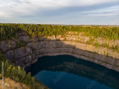 (Laufaufnahme, Drohne) Felswand eines Steinbruchs mit blauem See im Vordergrund