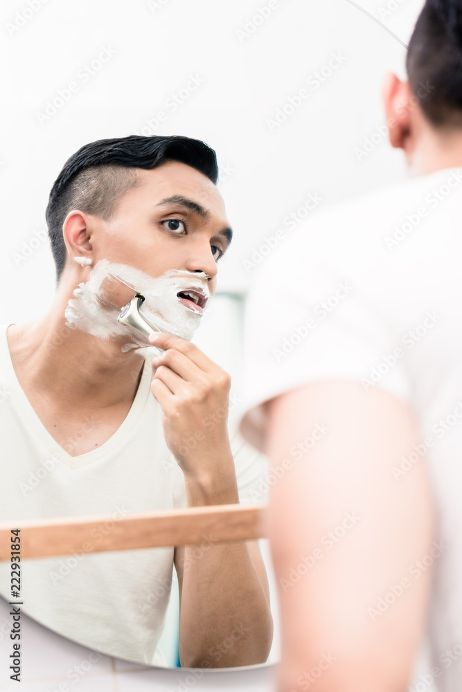 Young man looking in mirror shaving with razor