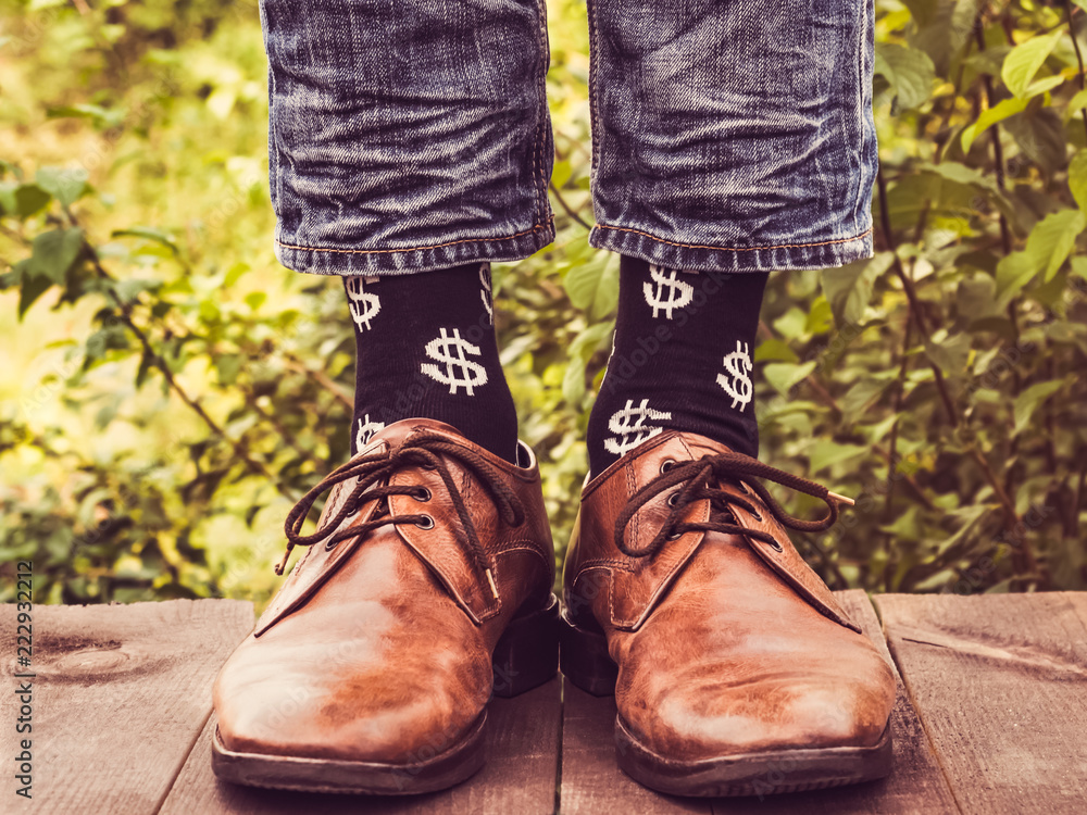 Men's legs in stylish shoes, black socks with patterns in the form of US dollars on a wooden terrace against the background of green trees. Beauty, fashion, elegance