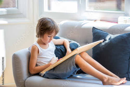 Prechool child, reading a book and playing with wooden educational toy at home, sitting on couch photo