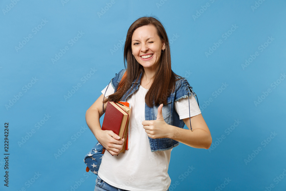 Portrait of young funny cute woman student in denim clothes with backpack blinking showing thumb up, hold school books isolated on blue background. Education in high school university college concept. Portrait of young funny cute woman student in denim clothes with backpack blinking showing thumb up, hold school books isolated on blue background. Education in high school university college concept.