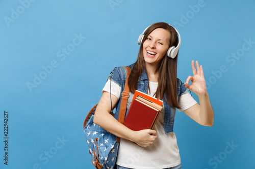 Quadro em tela Young cheerful woman student in denim clothes with backpack headphones listen music hold school books showing OK sign blinking isolated on blue background