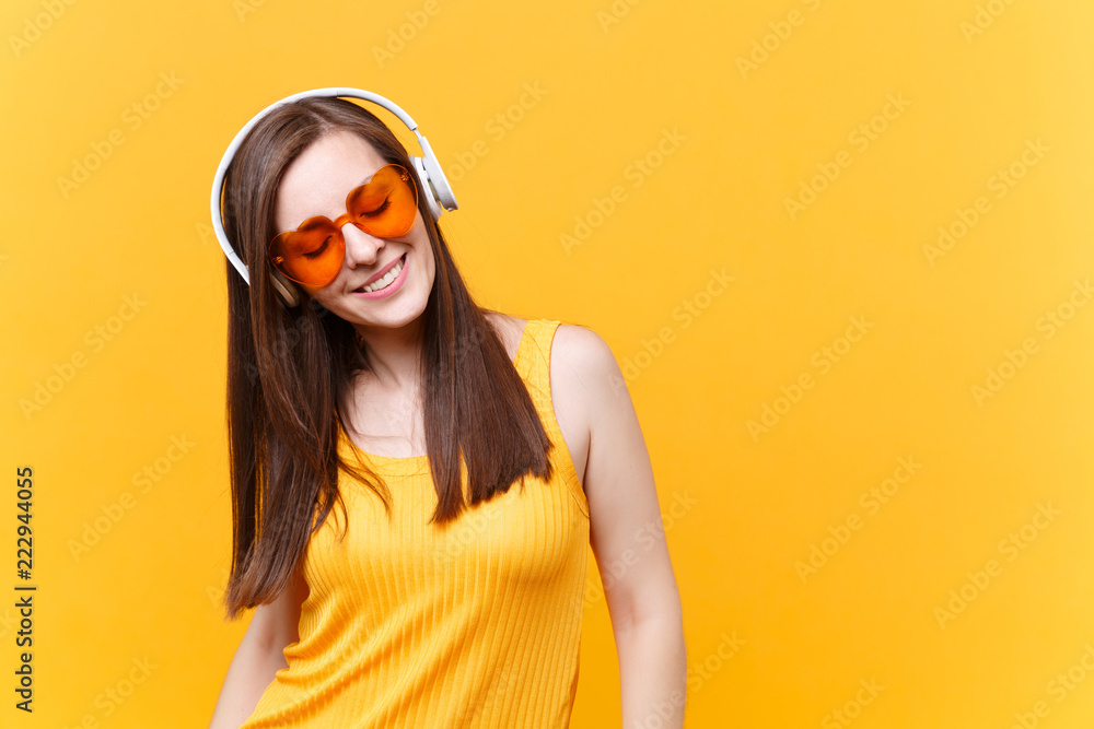 Portrait of calm smiling young woman in orange glasses listening music in headphones copy space isolated on yellow background. People sincere emotions, lifestyle concept. Advertising area. Rest, relax
