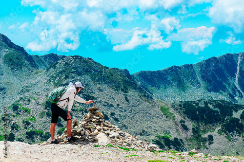 A young man hiker, reached the mountain top, building pyramid of rocks, beautiful alpine landscape, summertime.