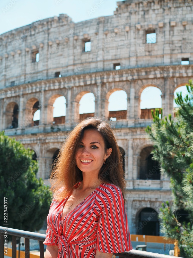 Brunette girl with long hair in a red striped dress stands on the background of the majestic ancient Colosseum in Rome