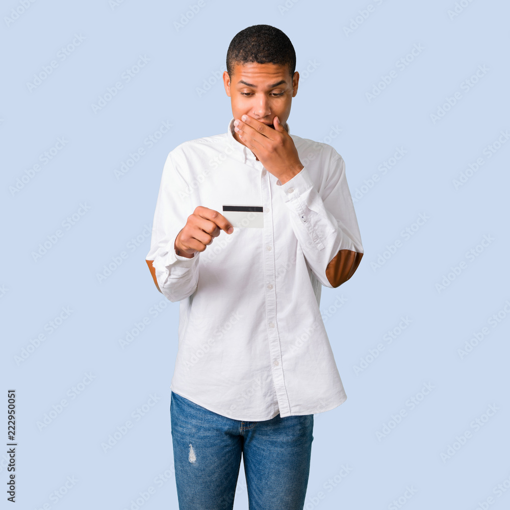 Young african american man with white shirt holding a credit card and surprised on isolated blue background Young african american man with white shirt holding a credit card and surprised on isolated blue background