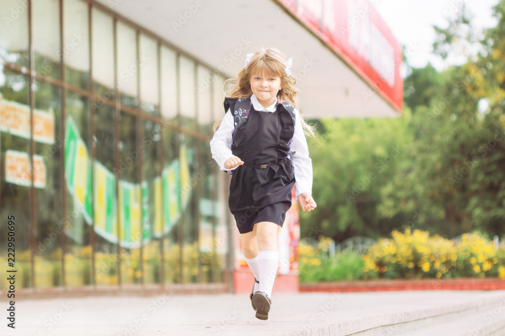 Little cheerful blue-eyed girl first-class with winded hair with bows in school uniform with a briefcase on September 1 runs jumping jolly at the school
