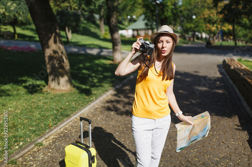 Young traveler tourist woman in hat with suitcase, city map take pictures on retro vintage photo camera in city outdoor. Girl traveling abroad to travel on weekend getaway. Tourism journey lifestyle.