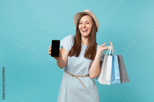 Portrait of woman in summer dress, straw hat holding packages bags with purchases after shopping, mobile cellphone with empty screen isolated on blue pastel background. Copy space for advertisement.