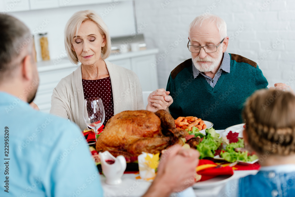 senior couple with family praying at served table with turkey before holiday dinner on thanksgiving