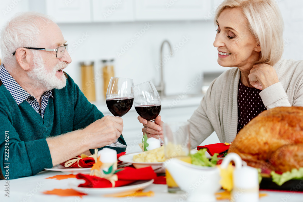 smiling senior couple clinking by wine glasses at serve table with baked turkey on thanksgiving smiling senior couple clinking by wine glasses at serve table with baked turkey on thanksgiving