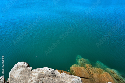 crystal clear blue cool water at the shore of a wild lake in a quarry without people with steep rocky beaches and large boulders on a summer warm day