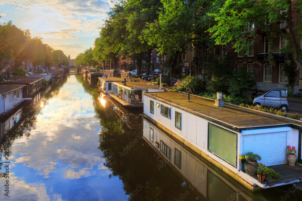 Fototapeta premium Łodzie mieszkalne przy kanałach światowego dziedzictwa UNESCO w Amsterdamie w Holandii w słoneczny letni poranek z błękitnym niebem i chmurami oraz lustrzanym odbiciem