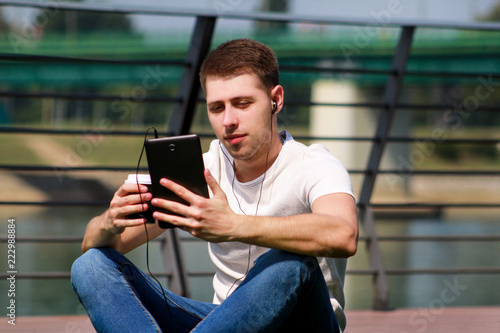 Handsome boy using tablet and headphones, drinking coffee to go, man sitting on grass and enjoying nice sunny day by river side. Model photo-shoot, urban fashion, lifestyle. White T-shirt and jeans.