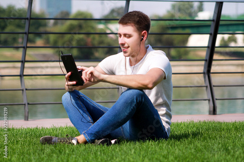 Handsome boy using tablet and headphones, drinking coffee to go, man sitting on grass and enjoying nice sunny day by river side. Model photo-shoot, urban fashion, lifestyle. White T-shirt and jeans.
