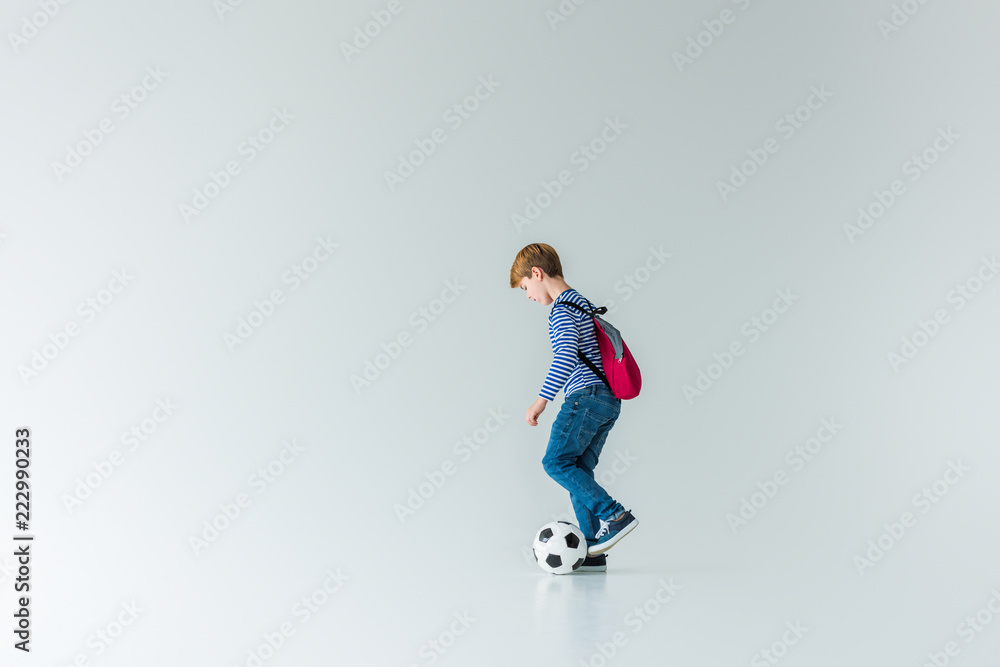 side view of schoolboy with backpack playing with fotball ball on white