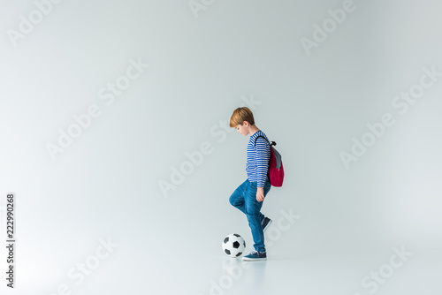 side view of adorable schoolboy with backpack playing with fotball ball on white