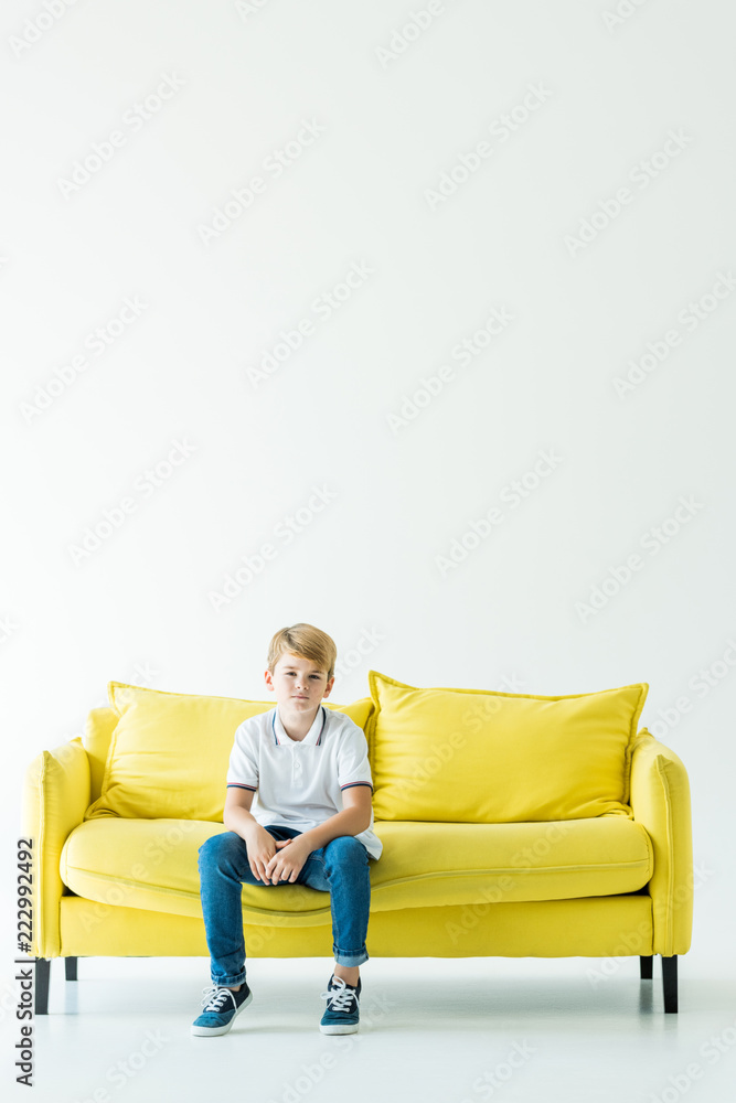 Obraz premium adorable boy sitting on yellow sofa and looking at camera on white