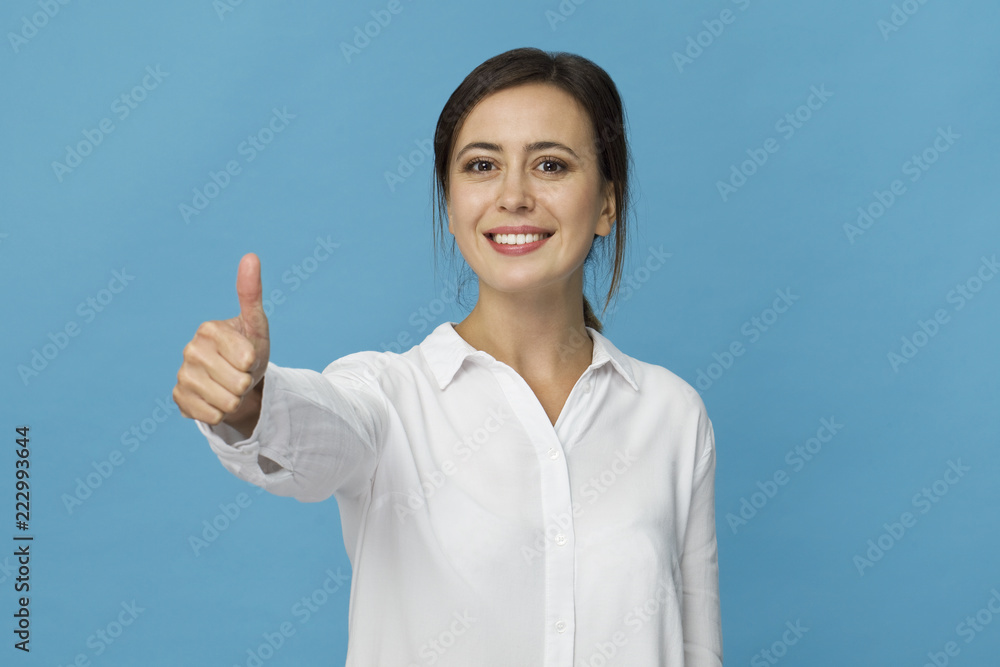 Smiling positive female with attractive look, wearing white T-shirt, posing against blue blank wall with OK gesture Smiling positive female with attractive look, wearing white T-shirt, posing against blue blank wall with OK gesture