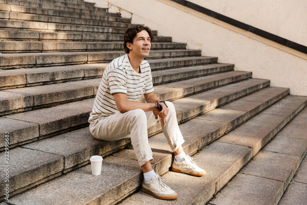 Happy young man sitting on steps outdoors looking aside holding phone. Happy young man sitting on steps outdoors looking aside holding phone.