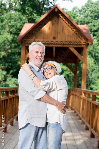 Hugging and smiling. Cheerful senior couple feeling good and smiling while hugging on the wooden bridge © Viacheslav Yakobchuk
