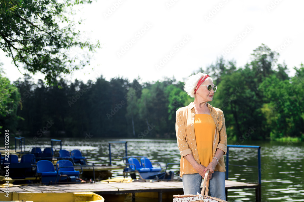 Ready for picnic. Relaxed senior woman standing near the river and smiling while holding a basket