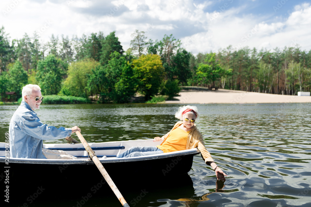 Touching water. Calm aged man rowing a little boat and his curious senior wife touching the water surface