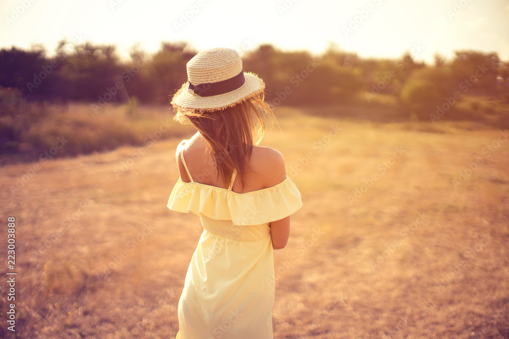 Back view of a beautiful young woman in yellow summer dress and straw hat having fun outdoor in countryside. Awesome warm summer day