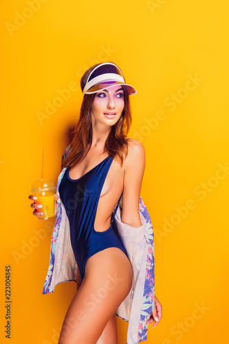 Side view of attractive young model in swimsuit and shirt enjoying tasty beverage and looking away while standing on yellow background © zamuruev