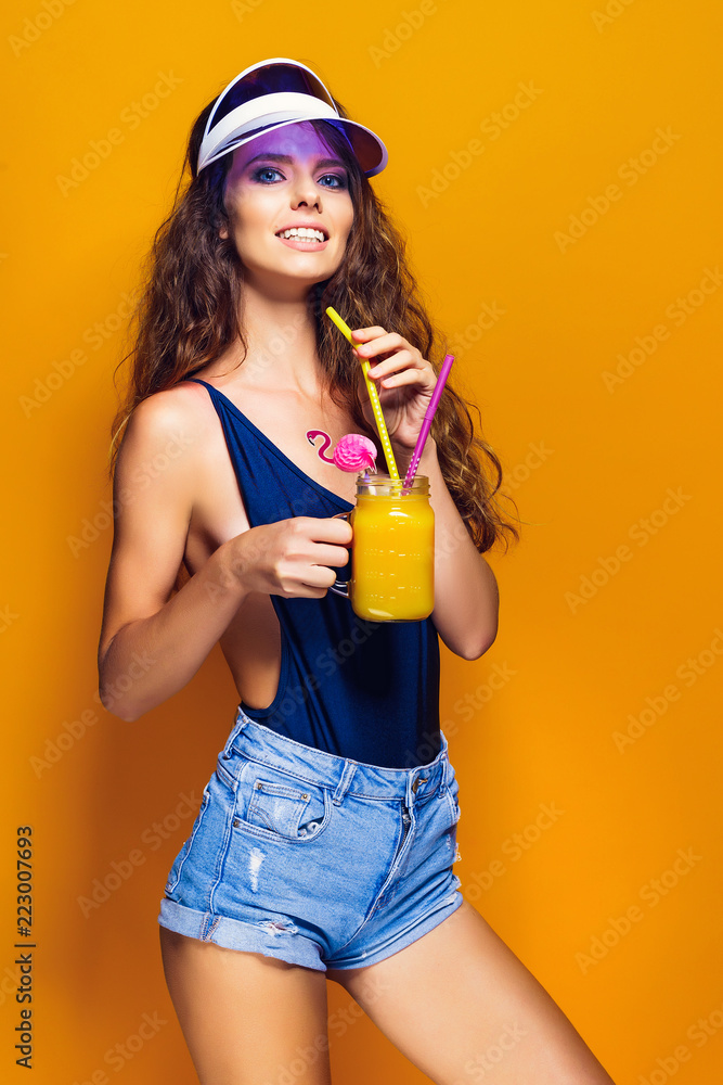 Sexy Woman in swimsuit and blue jeans shorts, trendy visor holding glass of fresh drink while standing on yellow, orange hot background Sexy Woman in swimsuit and blue jeans shorts, trendy visor holding glass of fresh drink while standing on yellow, orange hot background