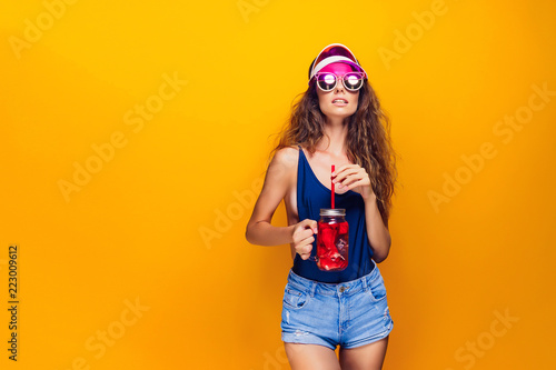 Sensual young woman in summer outfit, cap, sunglasses holding jar with fresh beverage and while standing on bright yellow background