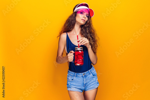 Sensual young woman in summer outfit holding jar with fresh beverage and while standing on bright yellow background