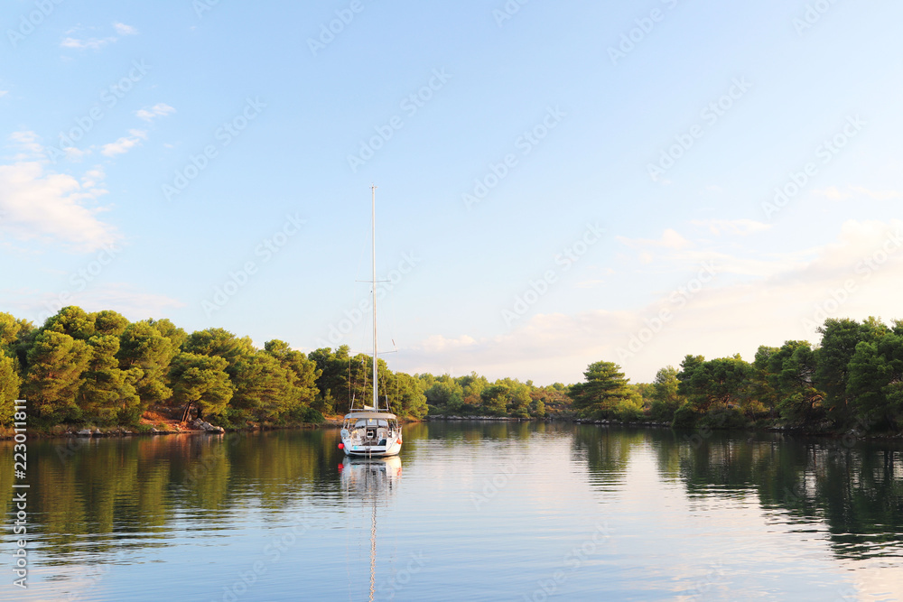 A sailing yacht is anchored in a picturesque bay surrounded by greenery of a coniferous forest. Calm and rest after sea crossing. Croatian Riviera on the Adriatic Sea of the Mediterranean area. A sailing yacht is anchored in a picturesque bay surrounded by greenery of a coniferous forest. Calm and rest after sea crossing. Croatian Riviera on the Adriatic Sea of the Mediterranean area.