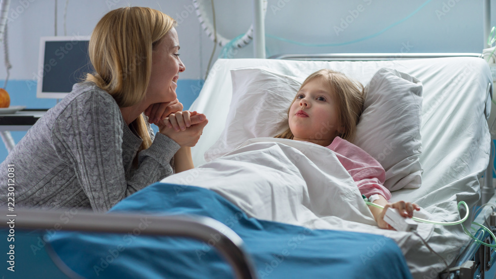 Cute Little Girl Lies on a Bed in the Children's Hospital, Her Mother Sits Beside, They Talk. Modern Pediatric Ward. Cute Little Girl Lies on a Bed in the Children's Hospital, Her Mother Sits Beside, They Talk. Modern Pediatric Ward.