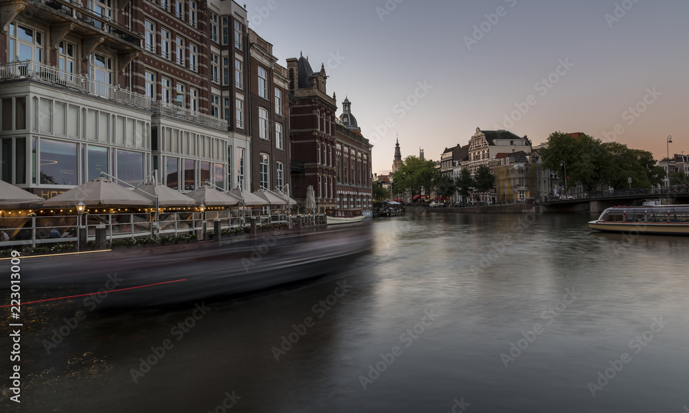 Amsterdam canal at dusk. A wide canal, with a moving canal boat sailing past the houses
