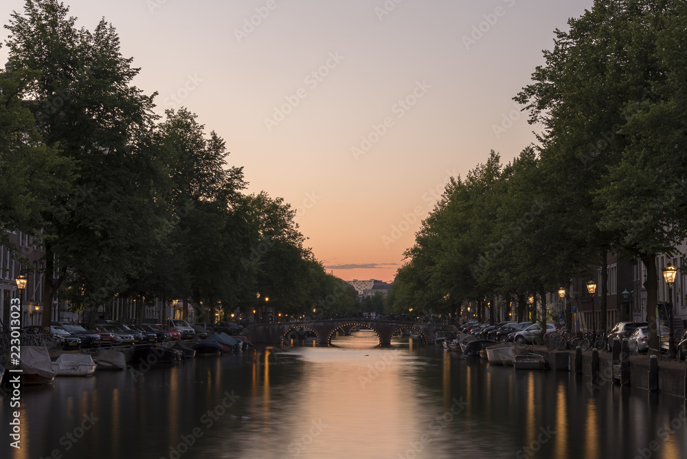 Sunset, looking down a canal in Amsterdam. The sky is orange and the street lights reflect on the calm water