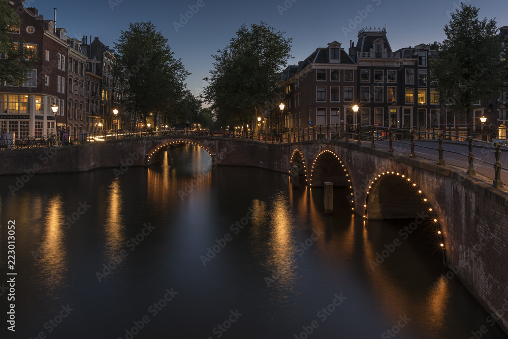Amsterdam canal and bridge, at dusk. The canal is calm and the lights around the bridges arches are reflecting on the smooth water.