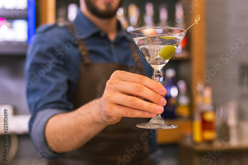 Tasty alcohol. Careful professional barman holding a glass of delicious alcohol with an olive